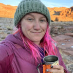 A woman with pink hair and a pink puffy yales a selfie in front of a washed out waterway with a desert background.